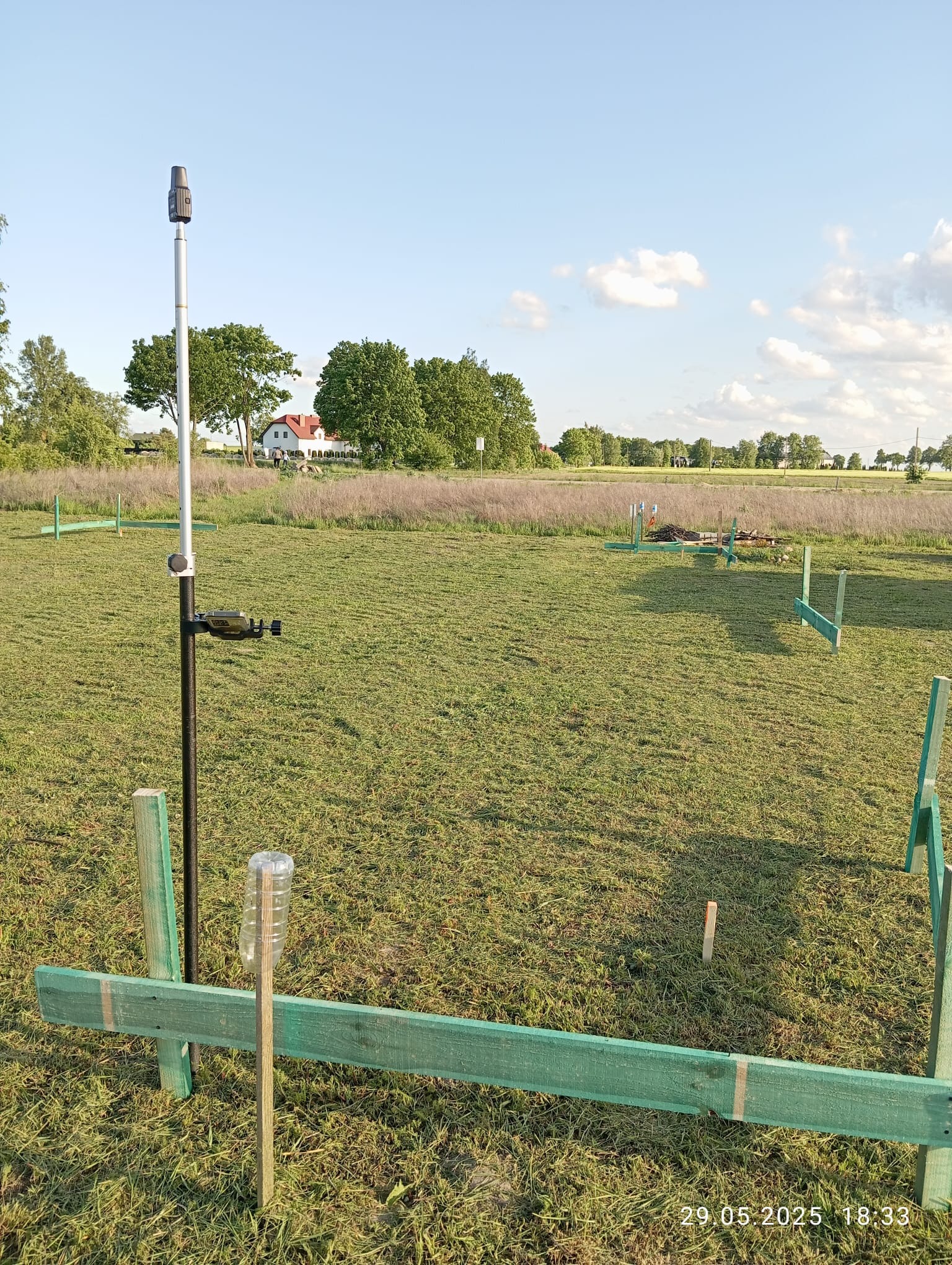 Pomiar geodezyjny na łące z użyciem niwelatora, widoczne drewniane znaczniki. W tle dom i drzewa pod błękitnym niebem.