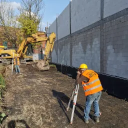Prace ziemne pod budowę drogi: operator koparki i geodeta dokonujący pomiarów niwelatorem na tle ściany z bloczków betonowych i budynku z czerwonej cegły.