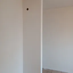 Świeżo pomalowane, jasne ściany w narożniku pokoju z widocznym otworem po gniazdku elektrycznym i fragmentem podłogi w trakcie remontu.