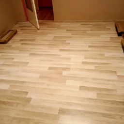 Jasne panele podłogowe ułożone w pokoju z otwartymi drzwiami, widoczne kartony z panelami przy ścianie.