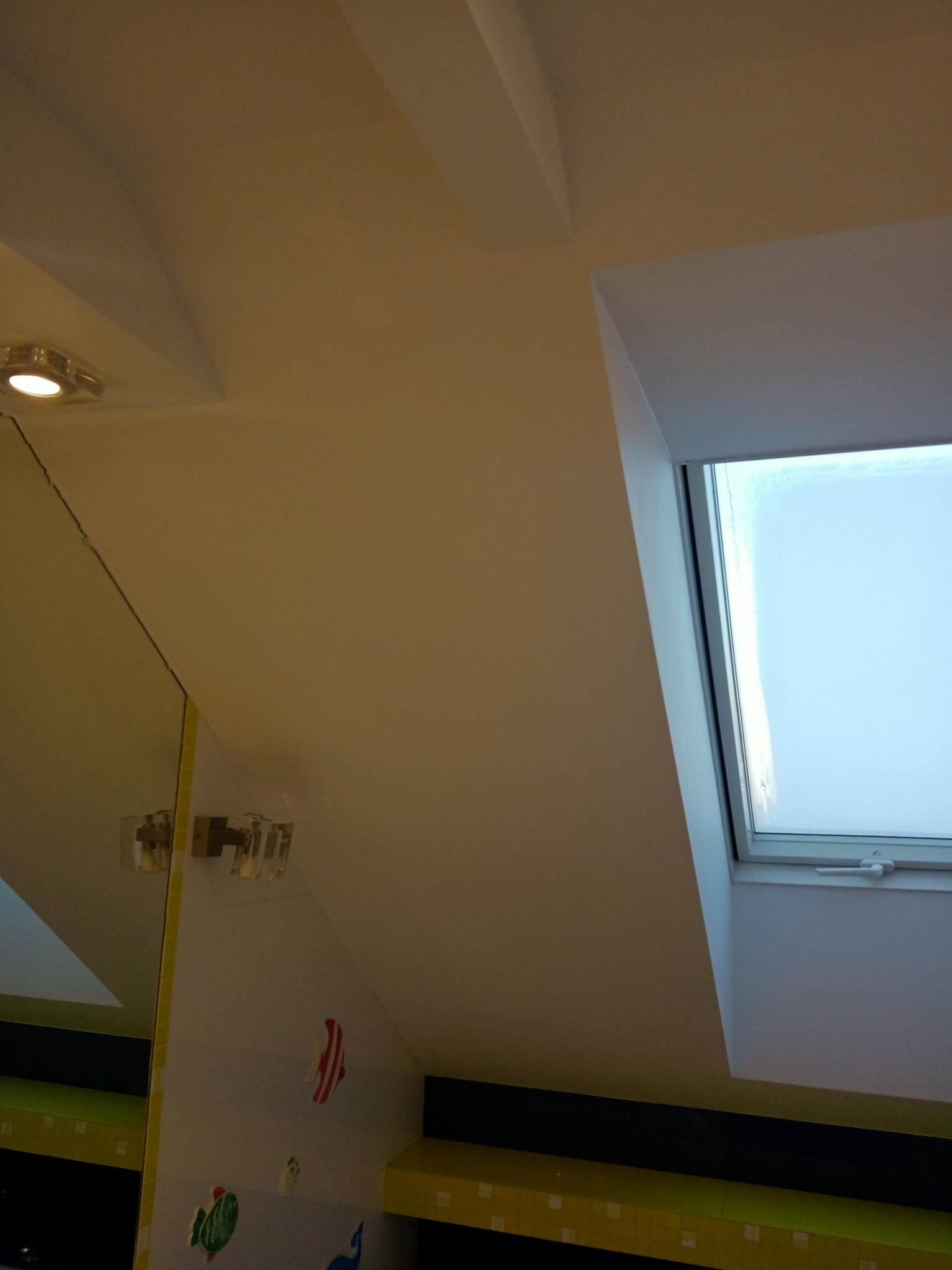 Poddasze łazienki po remoncie z oknem dachowym, lampą ścienną z przezroczystymi kloszami i lustrem z żółtą ramą, na ścianach naklejki z motywami morskimi.