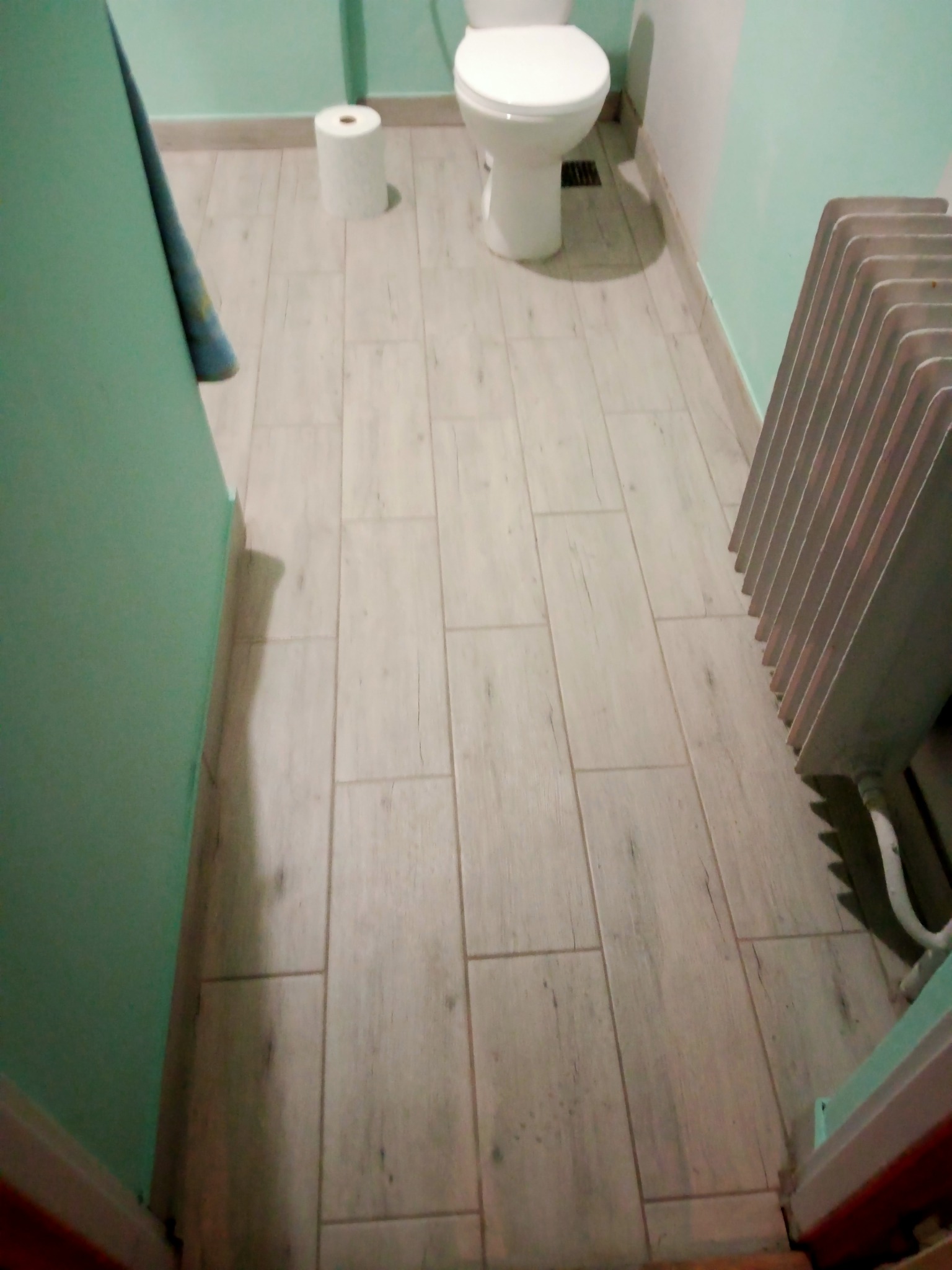 Jasne panele podłogowe imitujące drewno w łazience z toaletą i grzejnikiem żeberkowym, widoczny fragment turkusowej ściany i rolka papieru toaletowego.