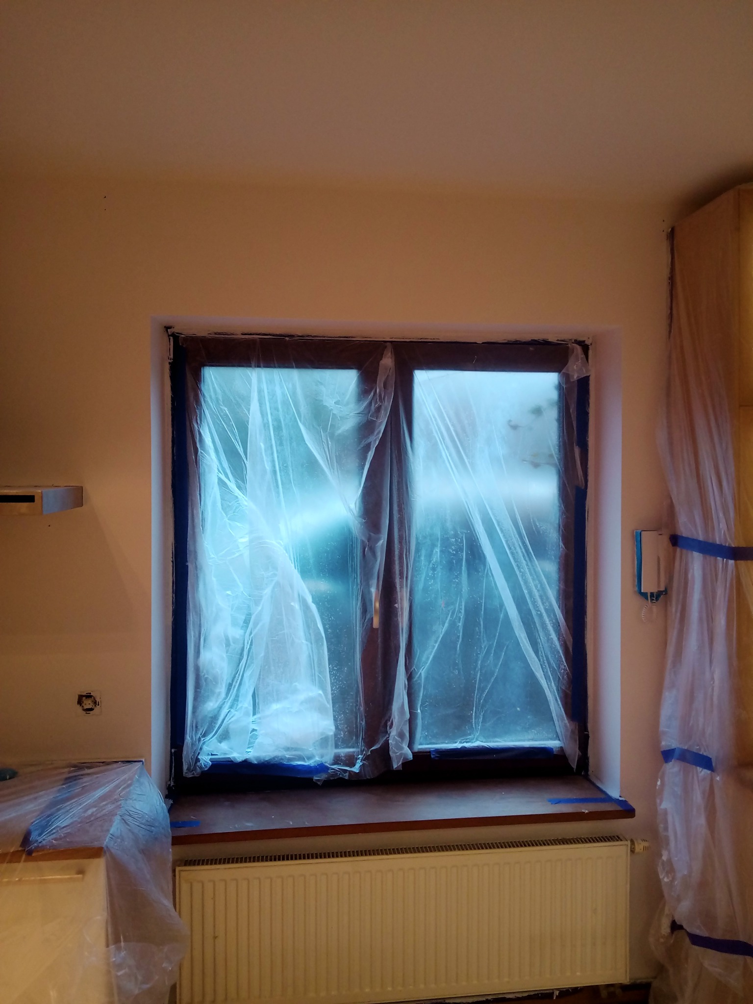 Okno zabezpieczone folią i taśmą malarską przed pracami wykończeniowymi, grzejnik i meble również osłonięte folią malarską, widoczne ślady po gniazdku elektrycznym.