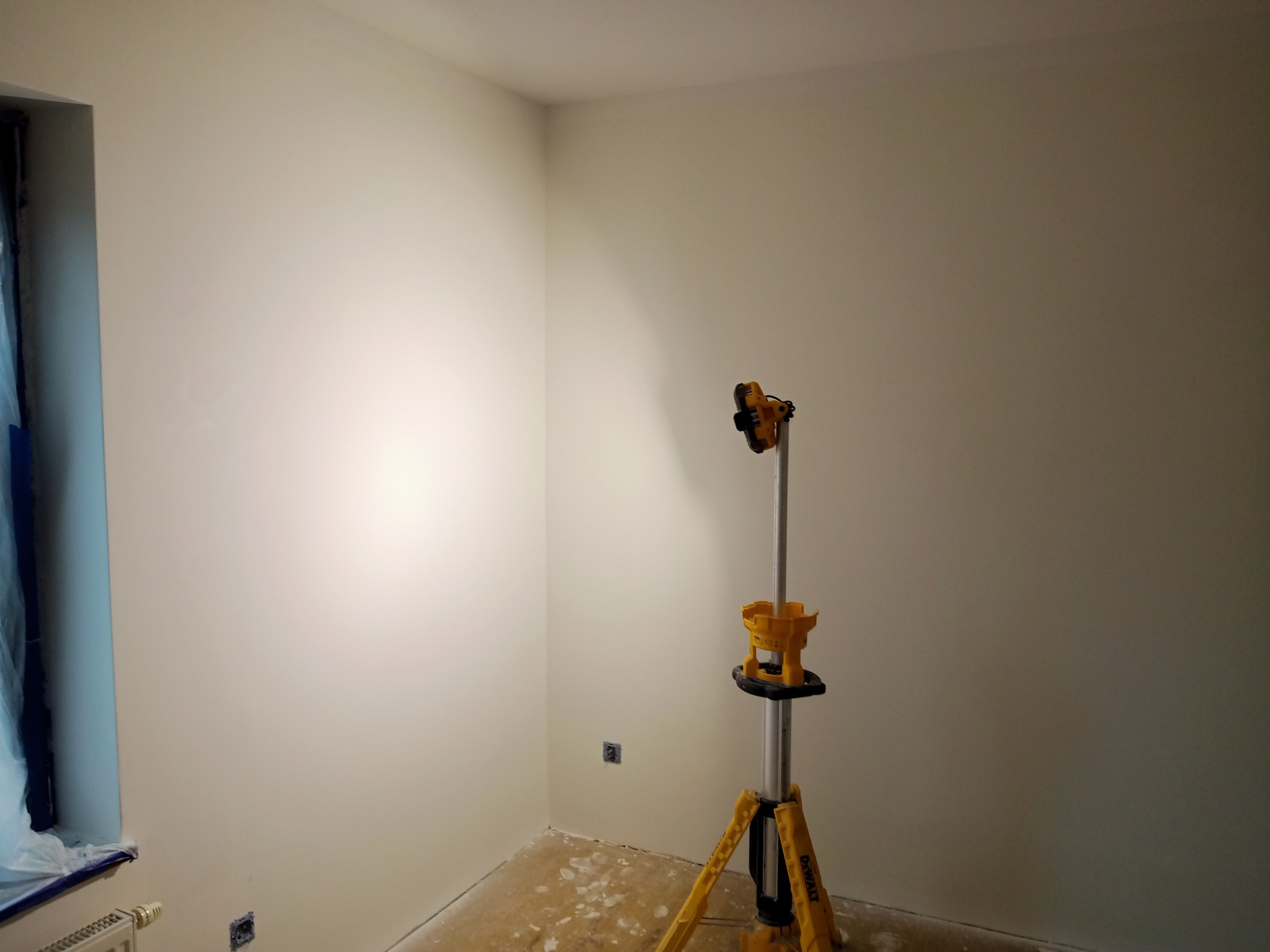 Pomalowane na biało ściany w rogu pokoju oświetlone lampą budowlaną DeWalt, okno zabezpieczone folią malarską.