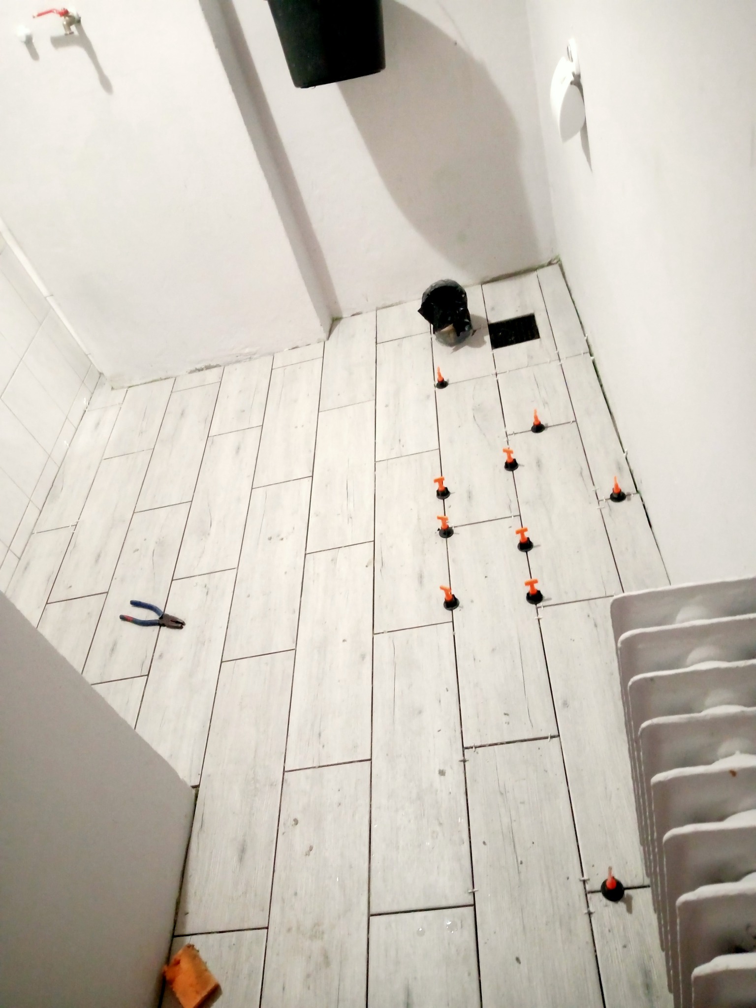 Układanie podłogowych płytek imitujących drewno w łazience, widoczne narzędzia glazurnicze - poziomujące klipsy i szczypce.