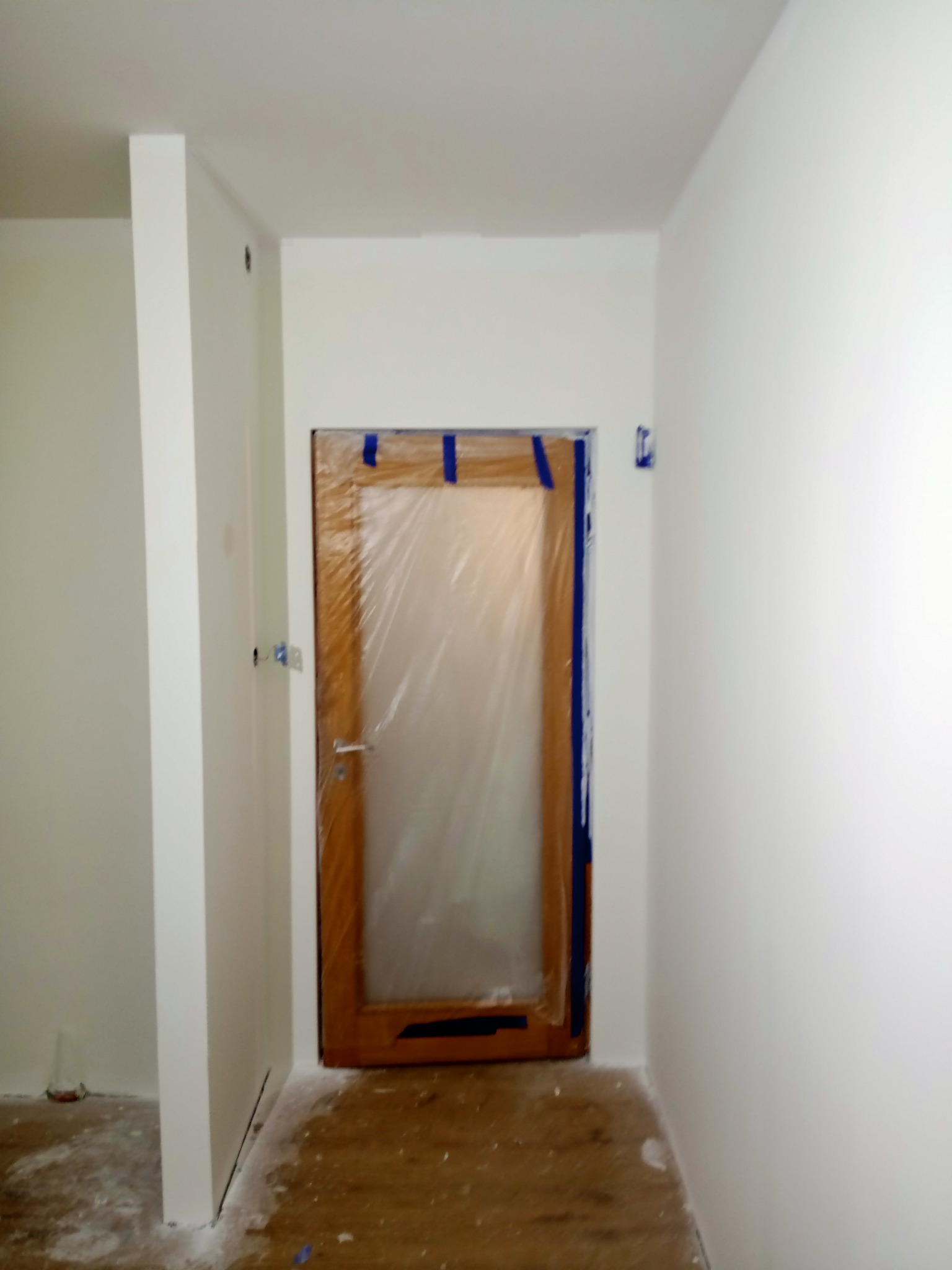Przygotowanie do malowania: drewniane drzwi zabezpieczone folią i niebieską taśmą malarską w białym pomieszczeniu z widocznymi śladami farby na podłodze.