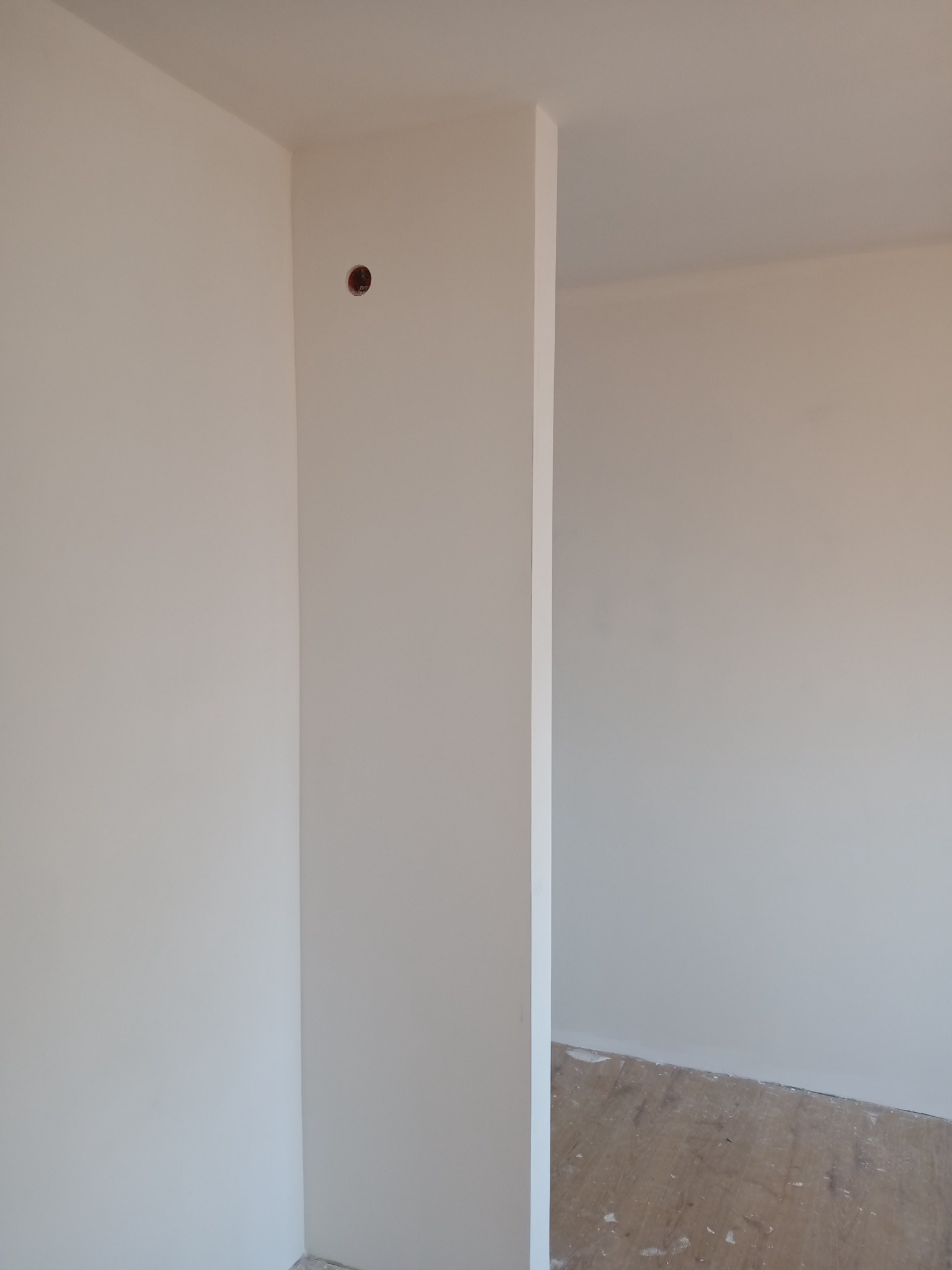 Świeżo pomalowane, jasne ściany w narożniku pokoju z widocznym otworem po gniazdku elektrycznym i fragmentem podłogi w trakcie remontu.