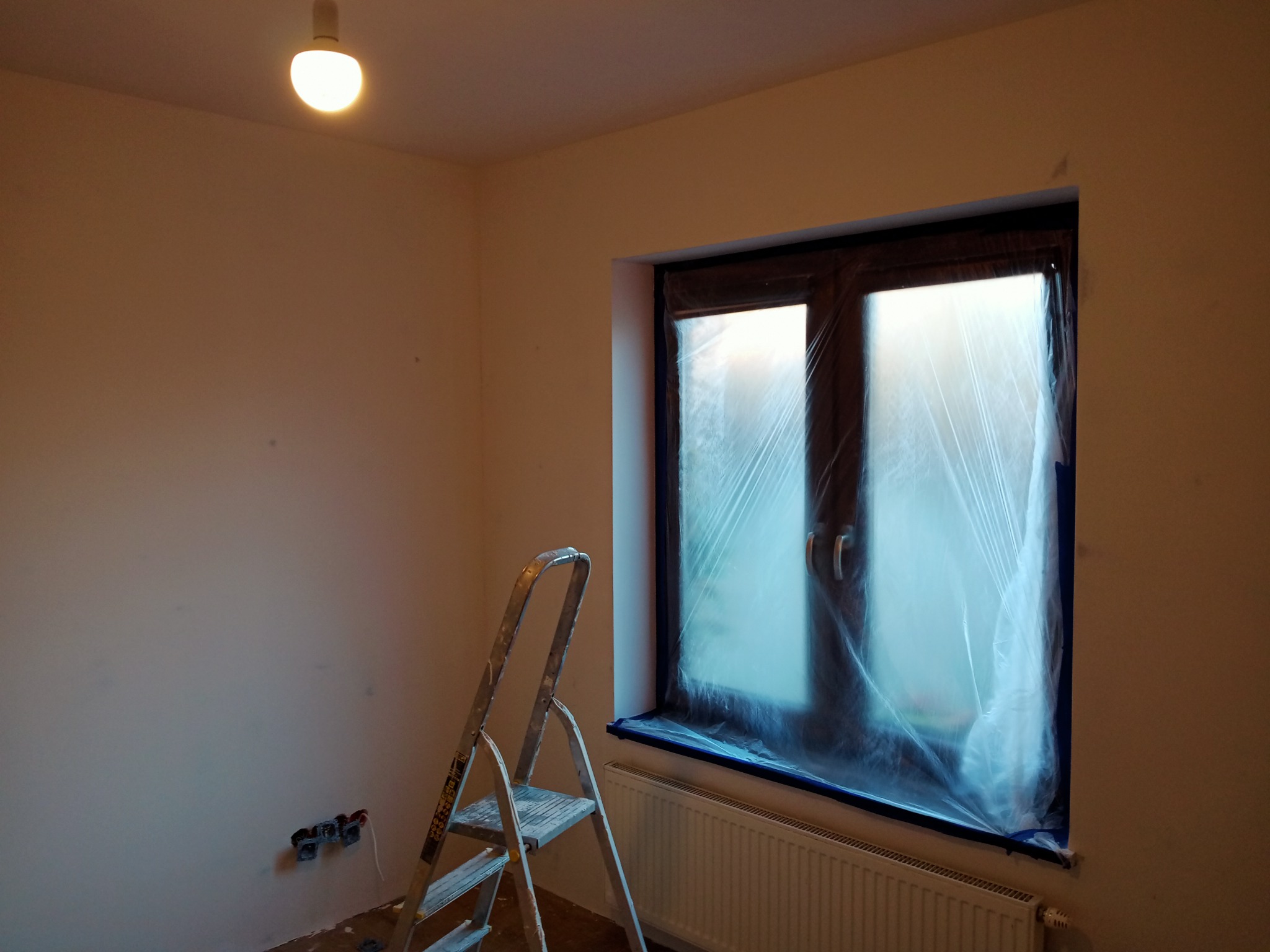 Pokój w trakcie przygotowań do malowania: okno zabezpieczone folią, drabina aluminiowa oparta o ścianę, widoczne gniazdka elektryczne i grzejnik.