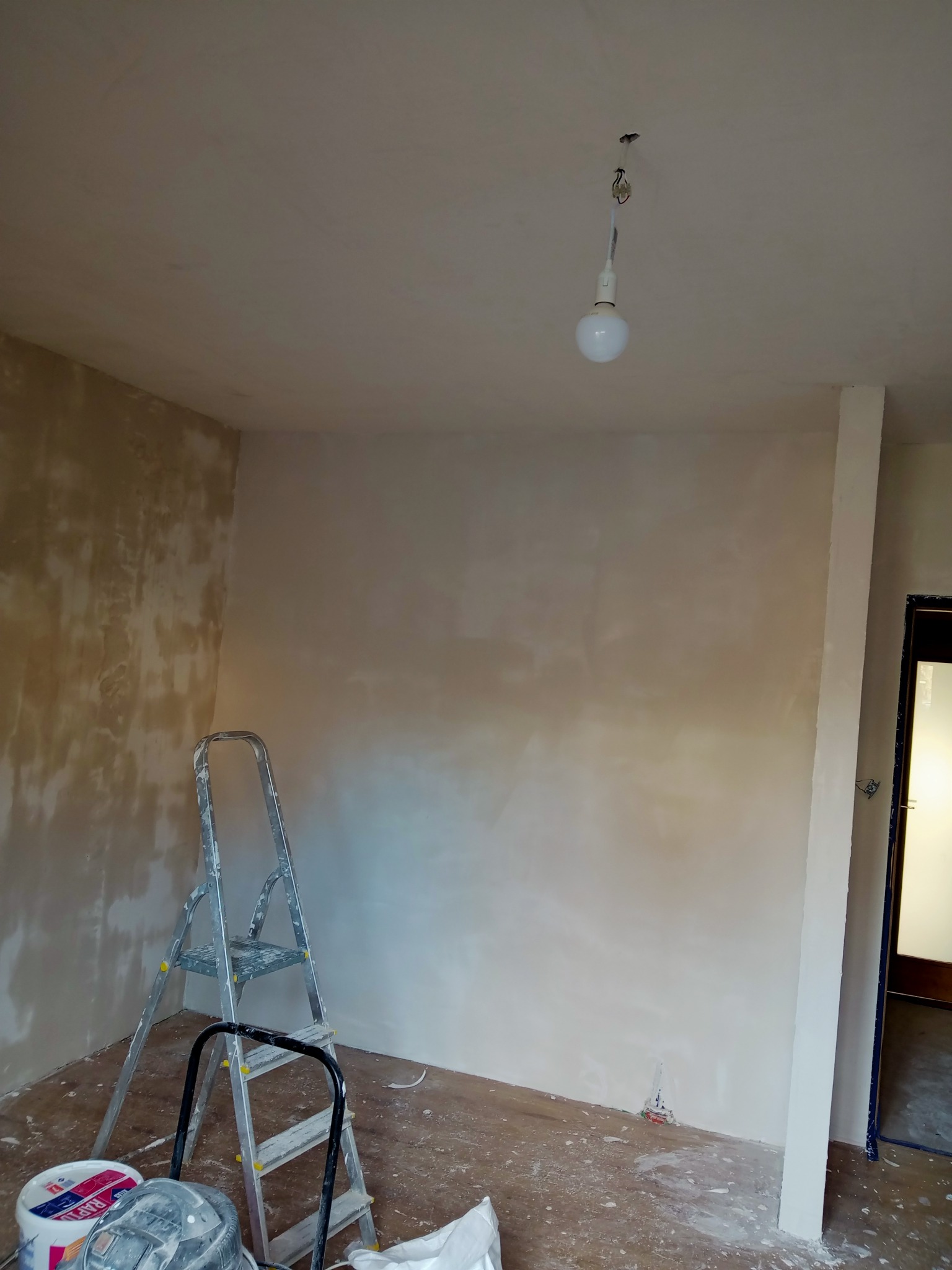 Pokój w trakcie wykańczania, ściany po nałożeniu gładzi, widoczna drabina malarska i wiadro z farbą, surowa żarówka zwisająca z sufitu.