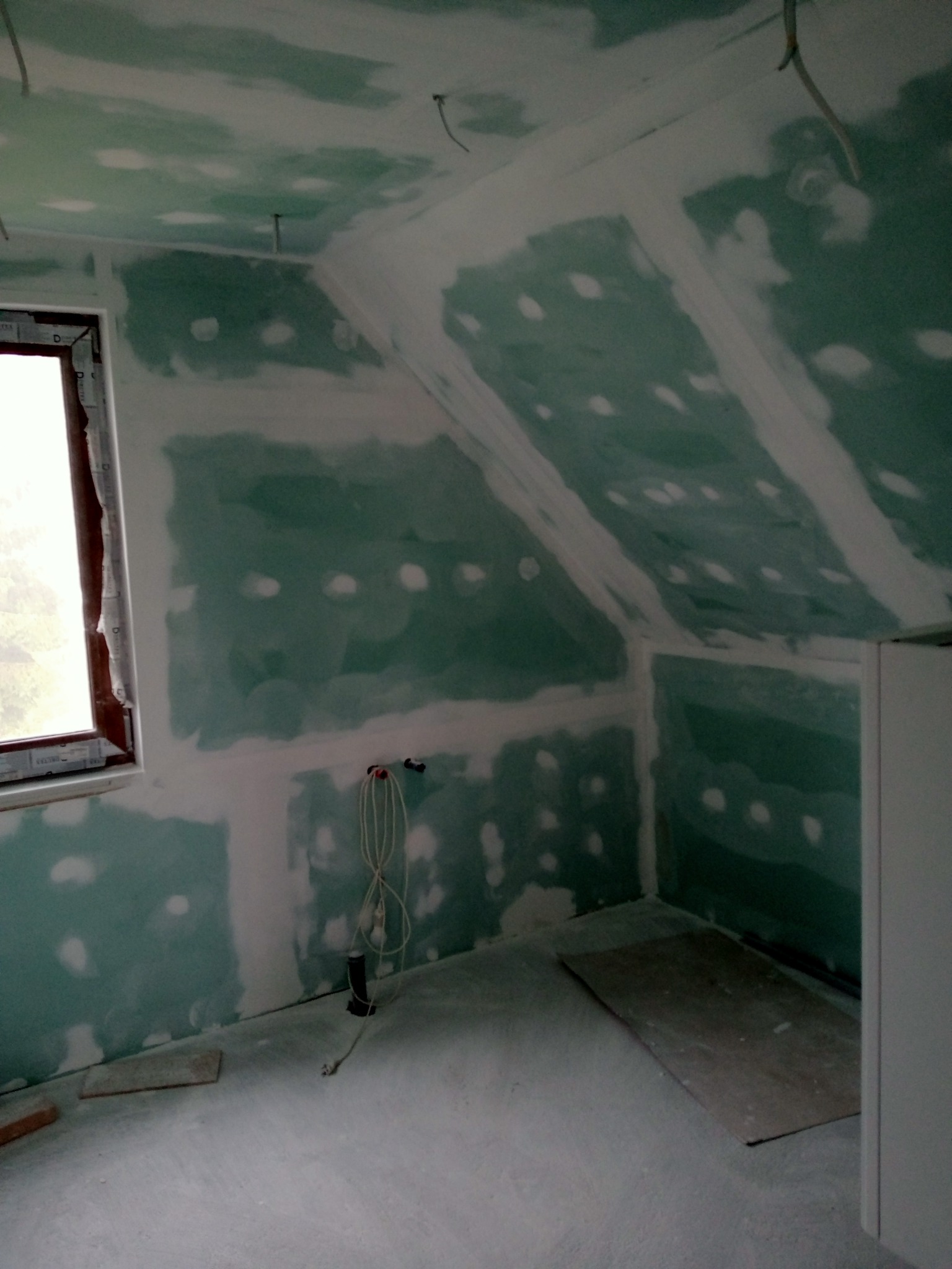 Poddasze z oknem, w trakcie montażu płyt kartonowo-gipsowych, widoczne łączenia szpachlowane na zielonych płytach, przewody elektryczne wystające ze ściany.