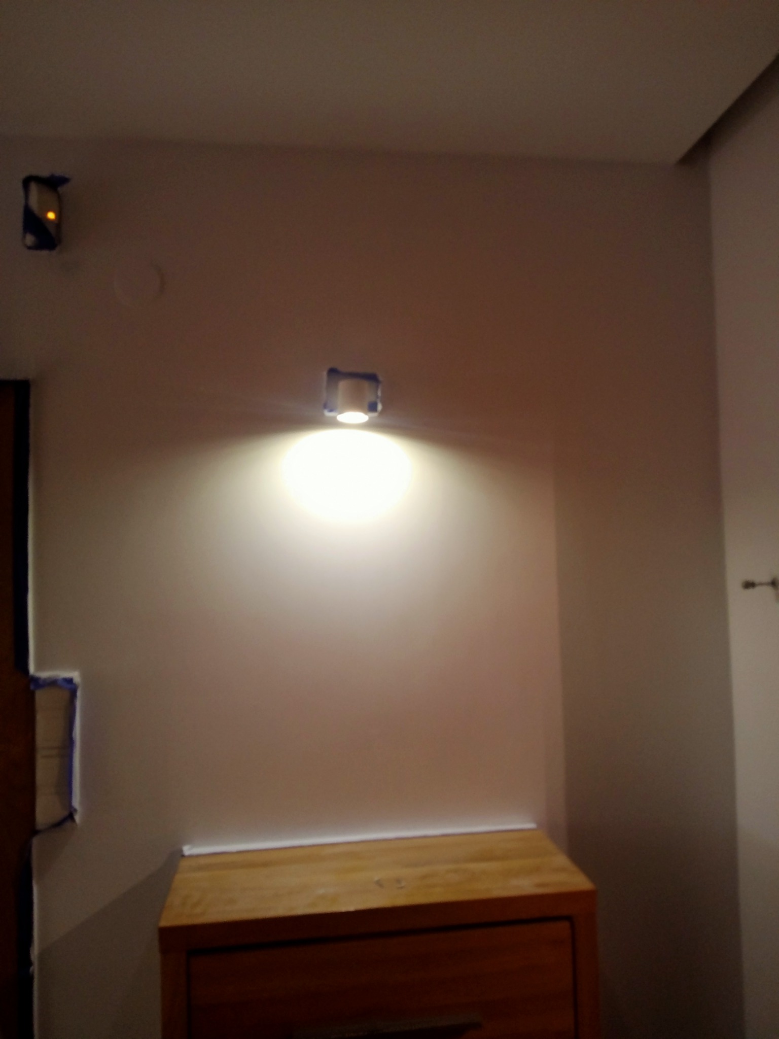 Ściana po malowaniu z zamontowanym kinkietem o ciepłej barwie światła, widoczna komoda z drewna.