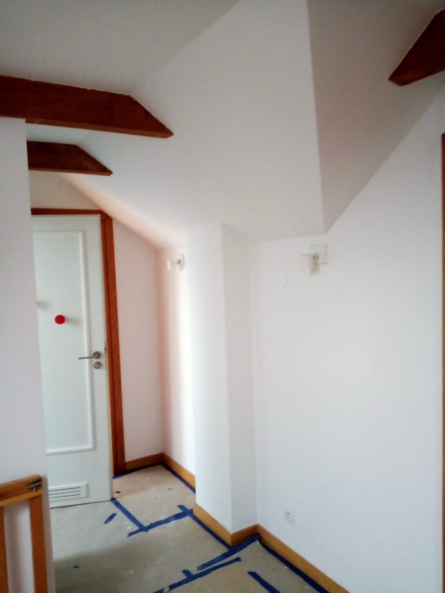 Korytarz w trakcie malowania, zabezpieczona podłoga taśmą malarską, świeżo pomalowane białe ściany z drewnianymi elementami wykończeniowymi i belkami na suficie.