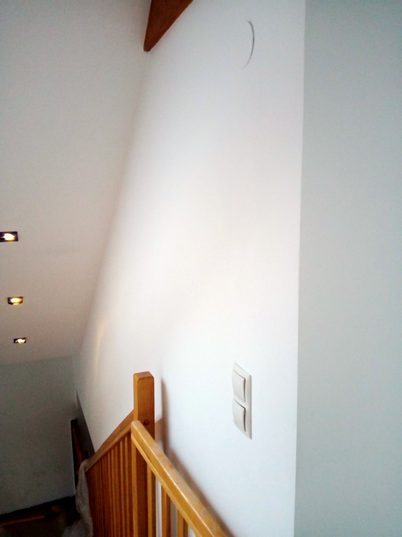 Świeżo pomalowana na biało ściana klatki schodowej z drewnianą balustradą, widoczne włączniki światła i sufit z wbudowanymi lampkami halogenowymi.