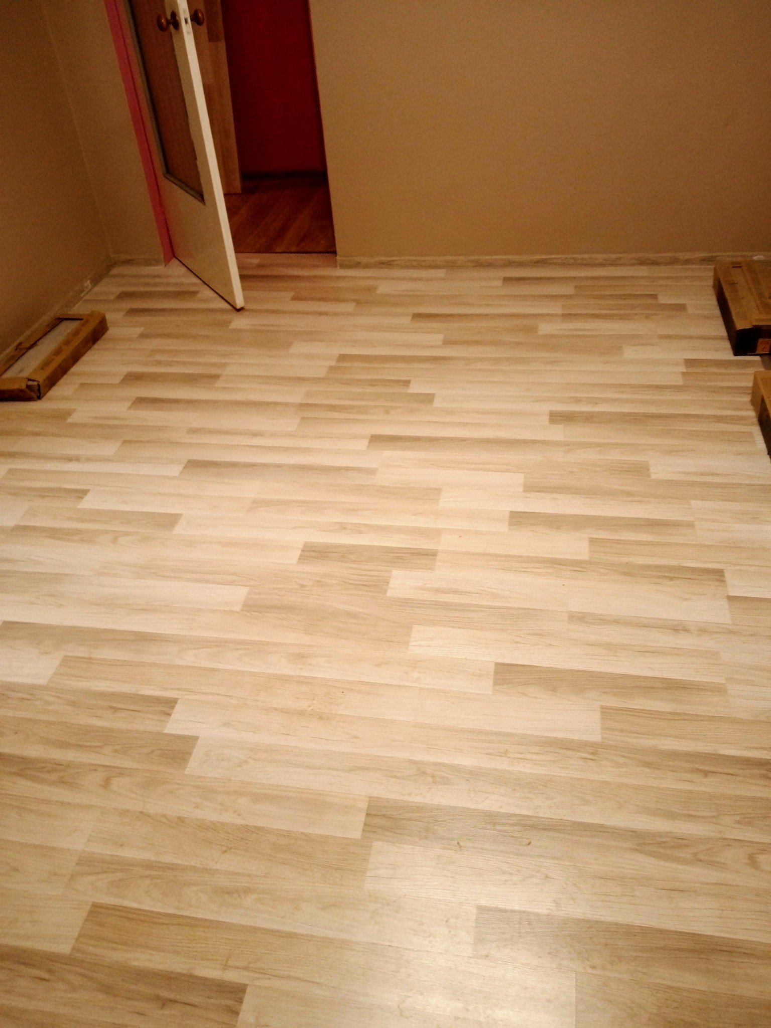 Jasne panele podłogowe ułożone w pokoju z otwartymi drzwiami, widoczne kartony z panelami przy ścianie.