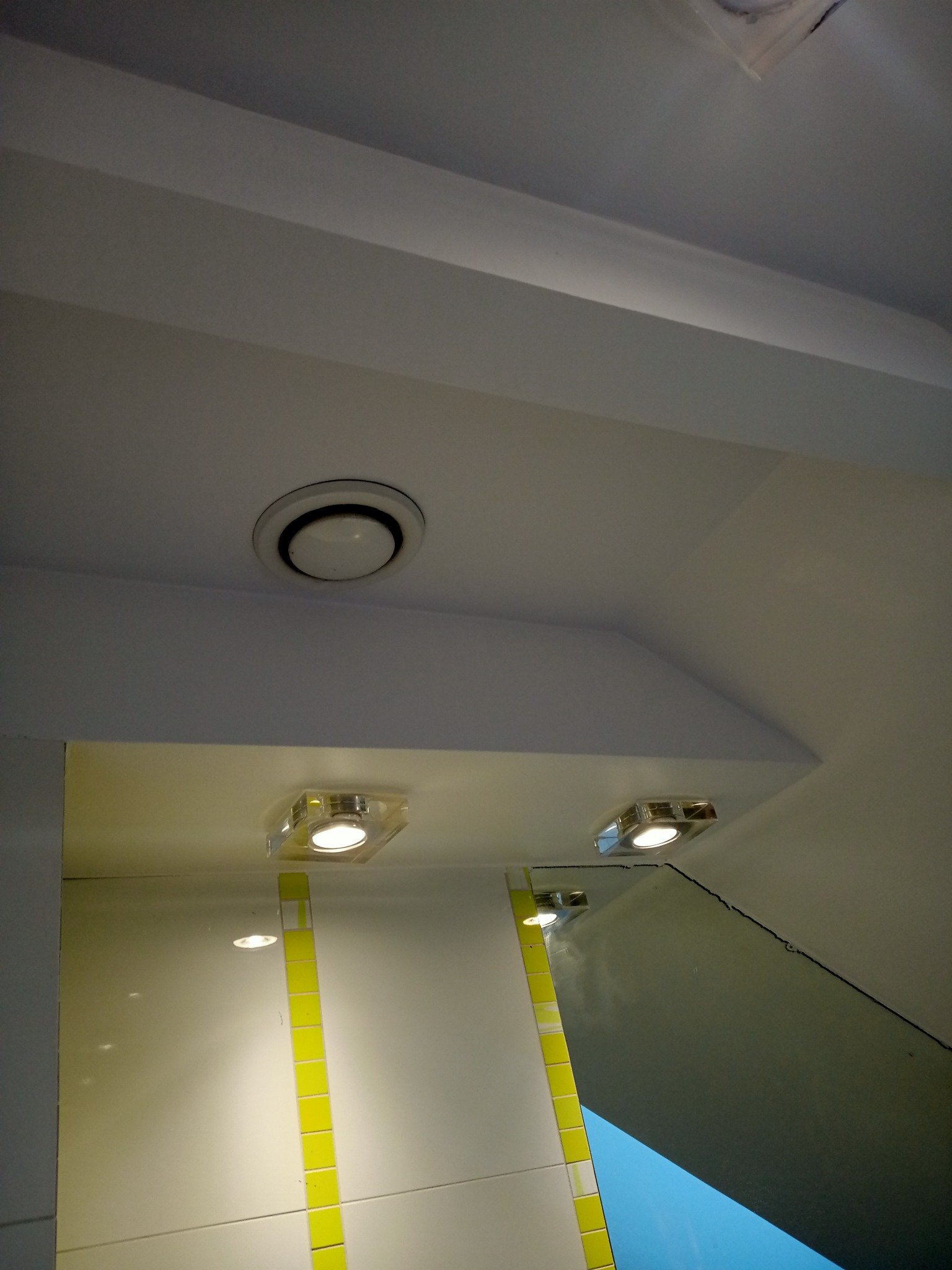Fragment łazienki w Proszowicach po remoncie: widoczny sufit z okrągłym nawiewnikiem, dwie halogenowe lampki w kwadratowych oprawach oświetlające białe płytki z pionowym paskiem żółtych płytek...