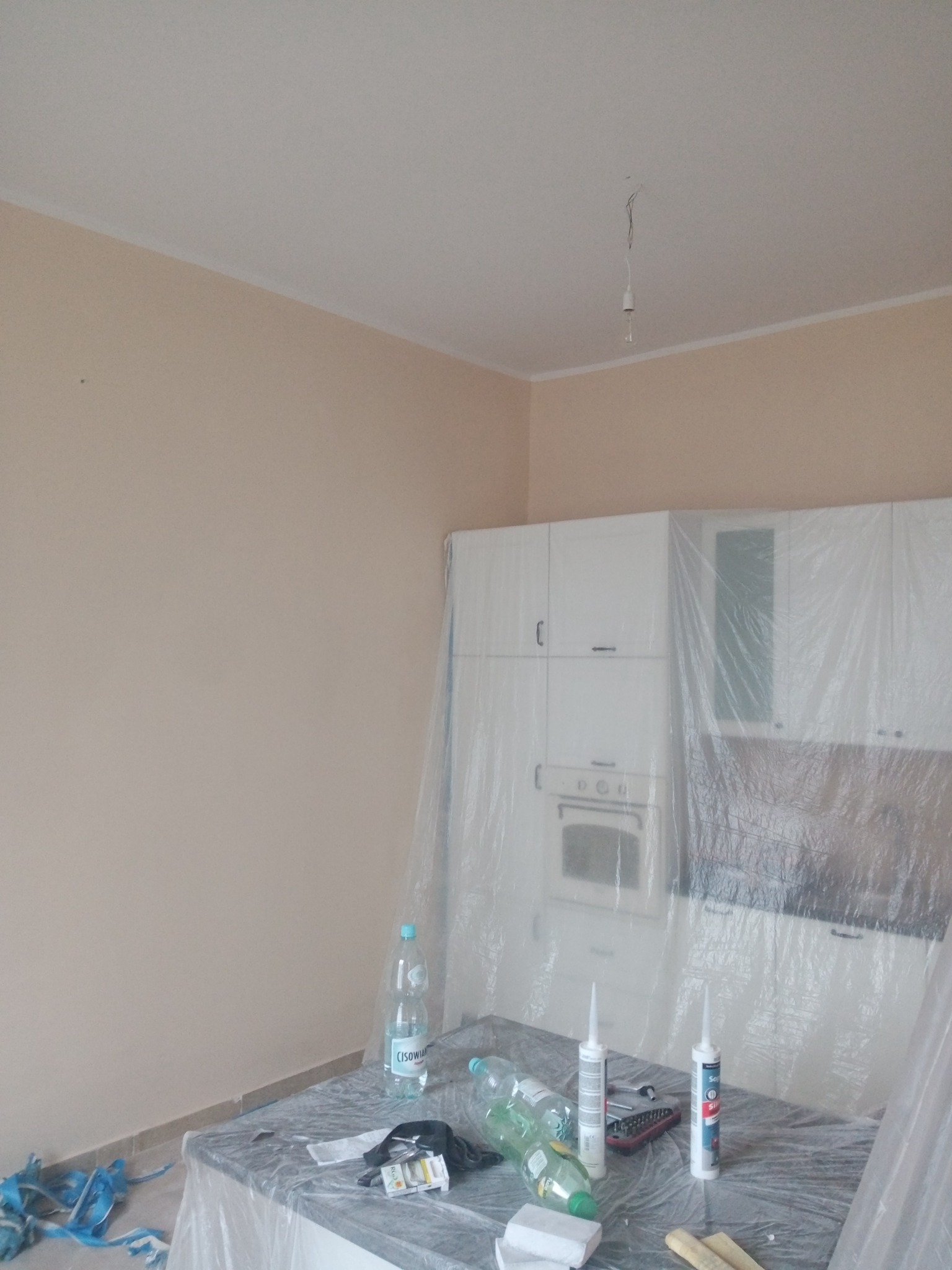 Przygotowania do remontu kuchni: ściany pomalowane na beżowo, meble kuchenne zabezpieczone folią ochronną, na blacie butelki z wodą, tuby silikonu i narzędzia.