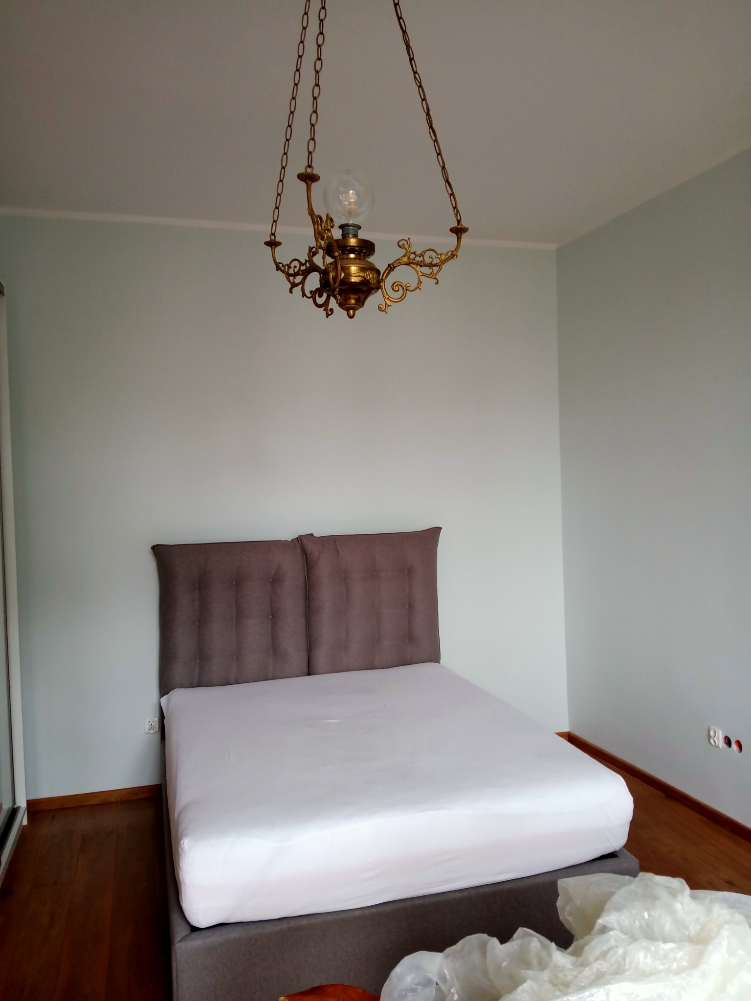Sypialnia z świeżo pomalowanymi ścianami w odcieniach szarości, na podłodze parkiet, a nad łóżkiem wisi ozdobny, złoty żyrandol.