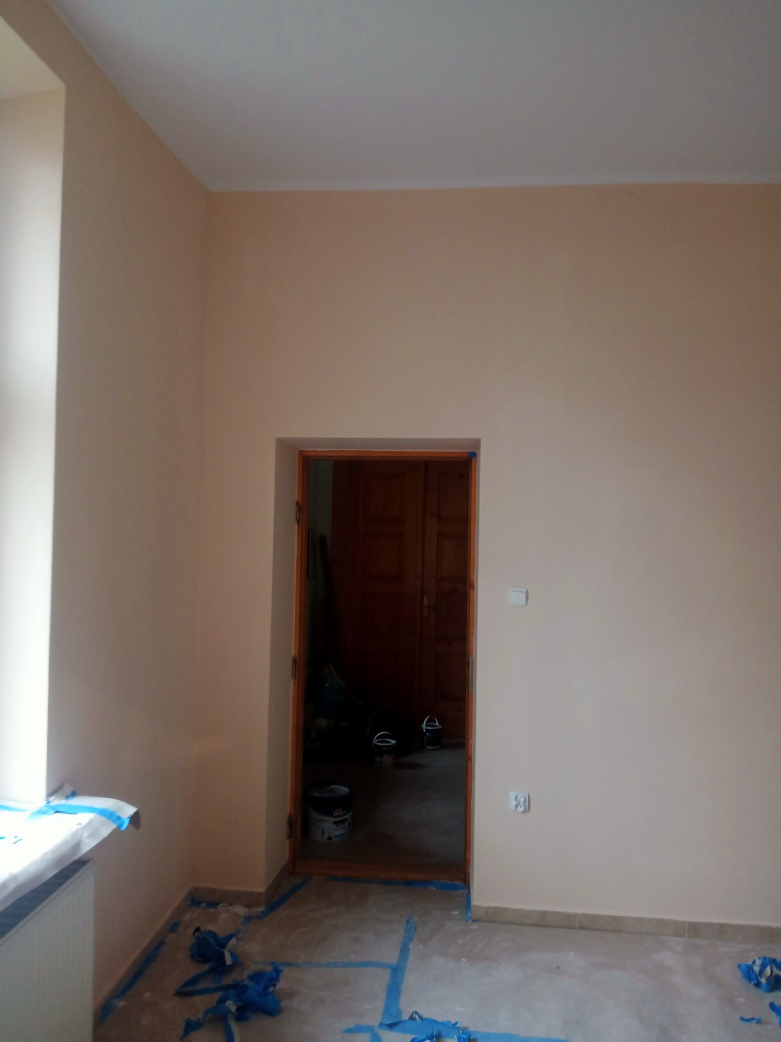 Świeżo pomalowane ściany na delikatny beż, widoczne zabezpieczenia folią malarską przy oknie i podłodze, otwarte drzwi do sąsiedniego pomieszczenia z widocznymi wiaderkami po farbie.