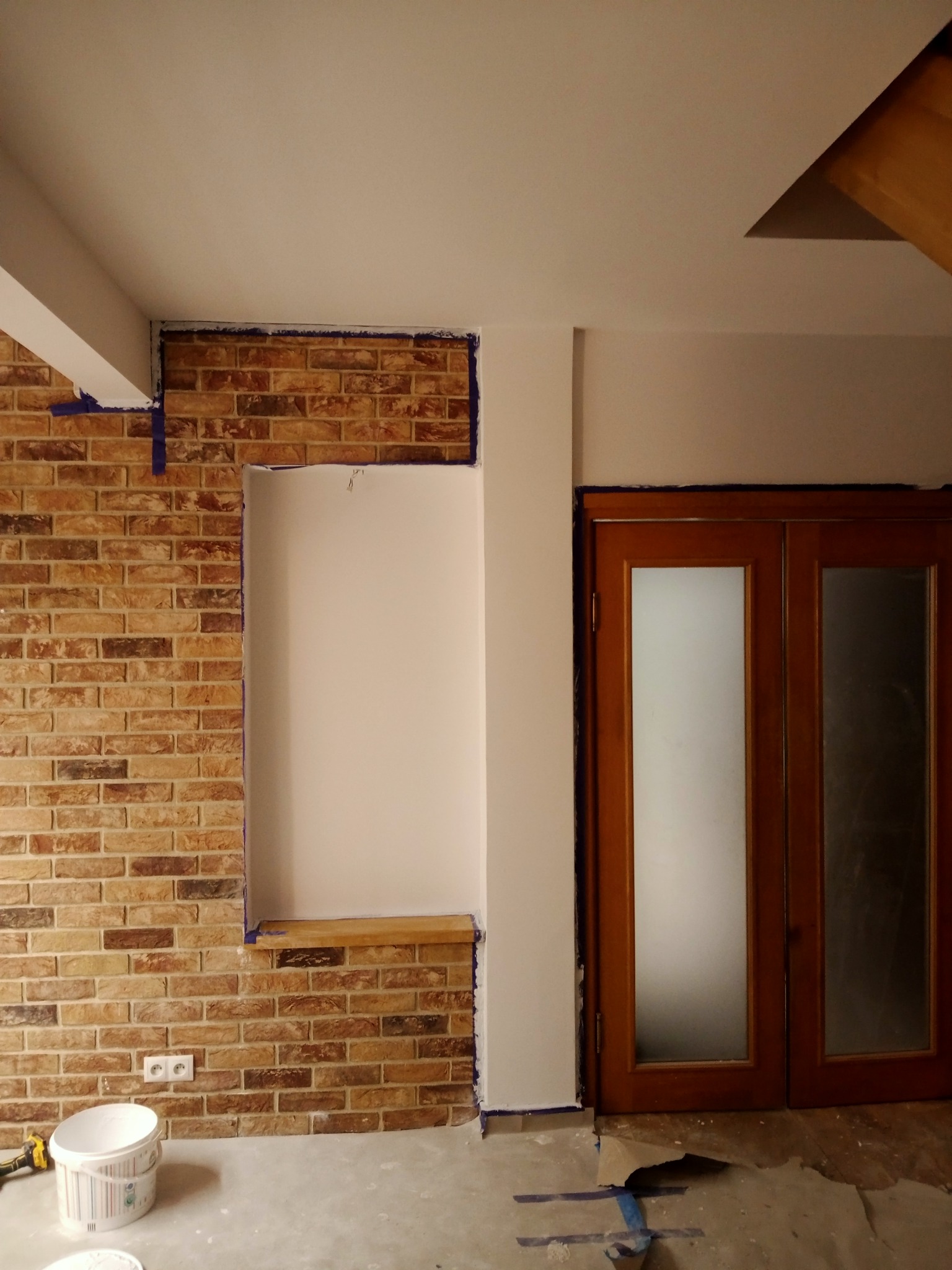 Fragment wnętrza w trakcie prac wykończeniowych: ściana z cegły klinkierowej z wydzielonym wnęką, zabezpieczona taśmą malarską, widoczne drzwi z przeszkleniami i wiadro z farbą.