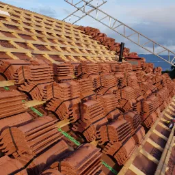 Czerwone dachówki ceramiczne ułożone w stosy na drewnianej konstrukcji dachu, zabezpieczone czarnymi taśmami; widoczna metalowa konstrukcja rusztowania i fragment krajobrazu w tle.