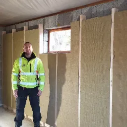 Pracownik w odblaskowej kurtce stoi w surowym pomieszczeniu z oknami, gdzie ściany z betonu komórkowego są częściowo ocieplone wełną mineralną przymocowaną do drewnianych łat.