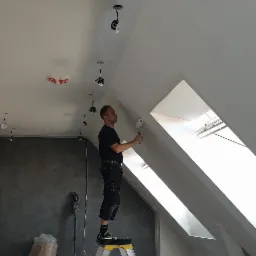 Pracownik na drabinie maluje białą farbą skośny sufit w pomieszczeniu ze świetlikami w Oświęcimiu, widoczne narzędzia malarskie i odkurzacz przemysłowy.