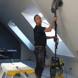 Pracownik szlifuje skośną ścianę poddasza za pomocą szlifierki z odkurzaczem, widoczna drabina z narzędziami i listwy przypodłogowe.