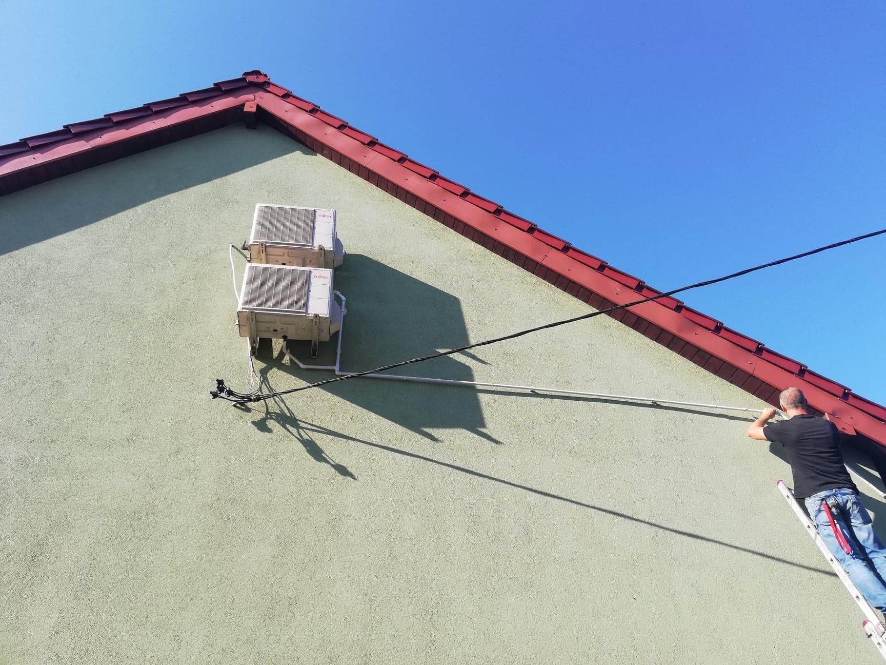 Dwa klimatyzatory Fujitsu zamontowane na ścianie szczytowej domu z czerwoną dachówką, instalator na drabinie montuje rurę odprowadzającą skropliny.