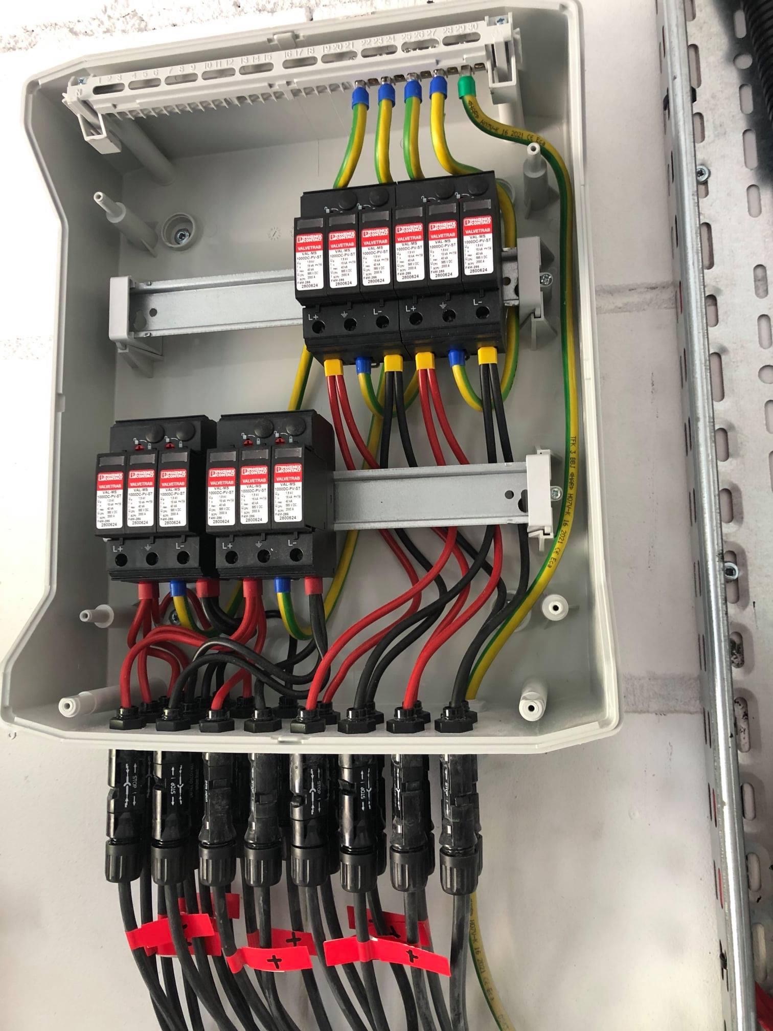 Otwarta skrzynka z zabezpieczeniami przeciwprzepięciowymi Phoenix Contact VALVETRAB dla instalacji fotowoltaicznej, z widocznymi przewodami oznaczonymi taśmami z symbolami plus i minus.