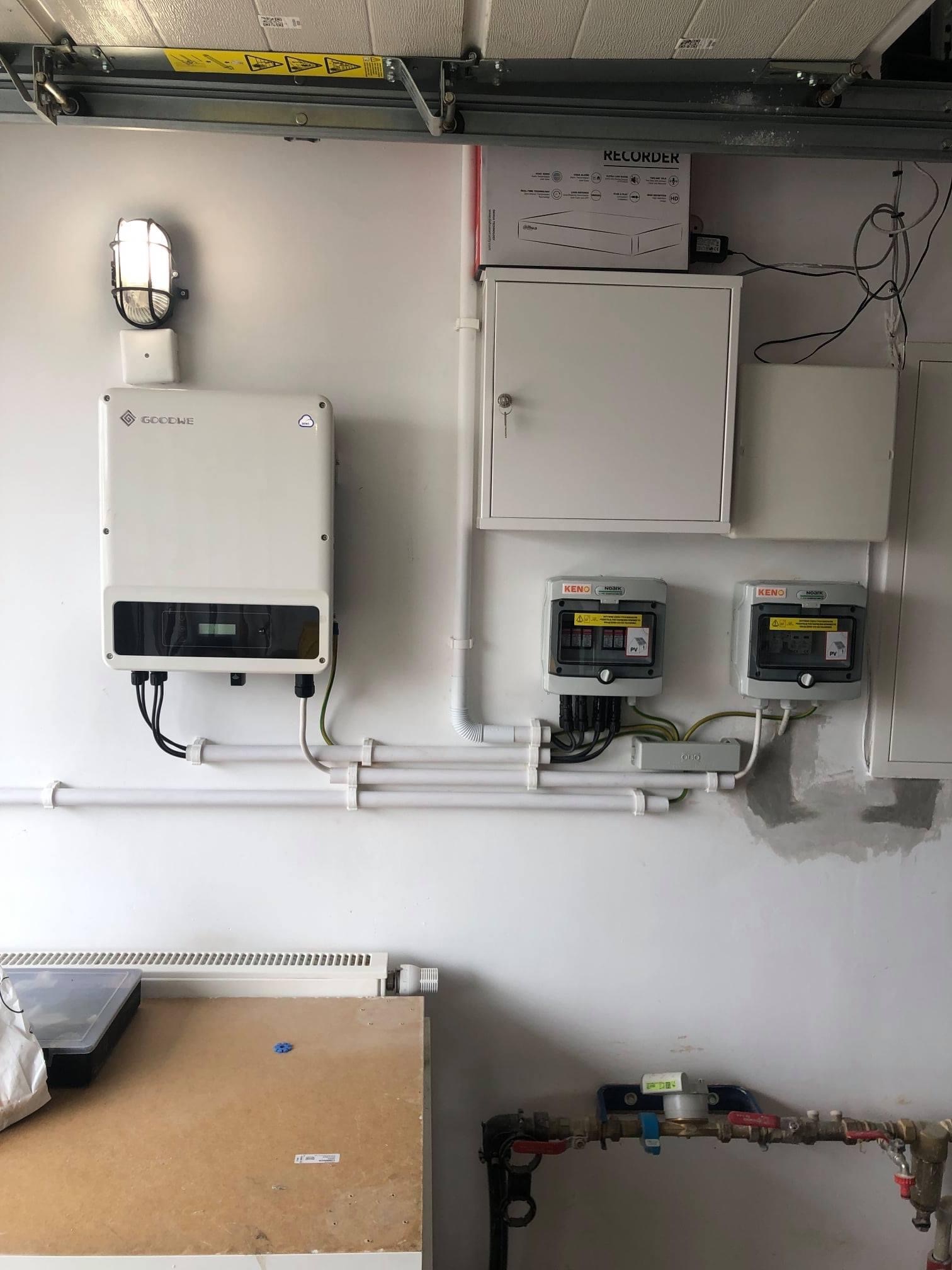 Biały falownik GOODWE zamontowany na ścianie w garażu obok lampy, z widocznymi przewodami, skrzynkami zabezpieczeń KENO, licznikiem wody i fragmentem warsztatu.