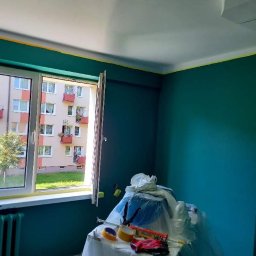 Sevma Art Sebastian Czerwinski - Pokój w trakcie malowania na ciemnozielono, z widocznymi narzędziami malarskimi na stole przykrytym folią, taśma malarska przy suficie i oknie, oraz widok na blok z balkonami za oknem.