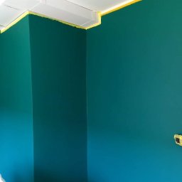 Sevma Art Sebastian Czerwinski - Ściana pomalowana na intensywny turkusowy kolor, zabezpieczona żółtą taśmą malarską przy białym suficie i narożniku, widoczne ślady farby na taśmie.