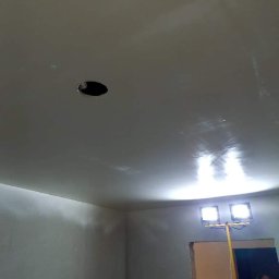 Sevma Art Sebastian Czerwinski - Gładki, świeżo pomalowany sufit z otworem na oświetlenie, oświetlony dwoma lampami halogenowymi na statywie, widoczny fragment ściany i fragment sylwetki osoby w tle.