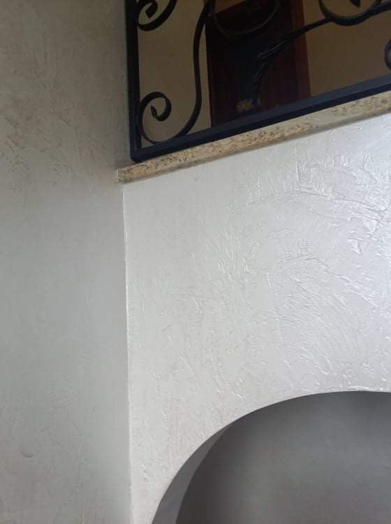 Jasna, teksturowana ściana w rogu pomieszczenia, wykończona ozdobnym, metalowym elementem nad łukowatym przejściem.