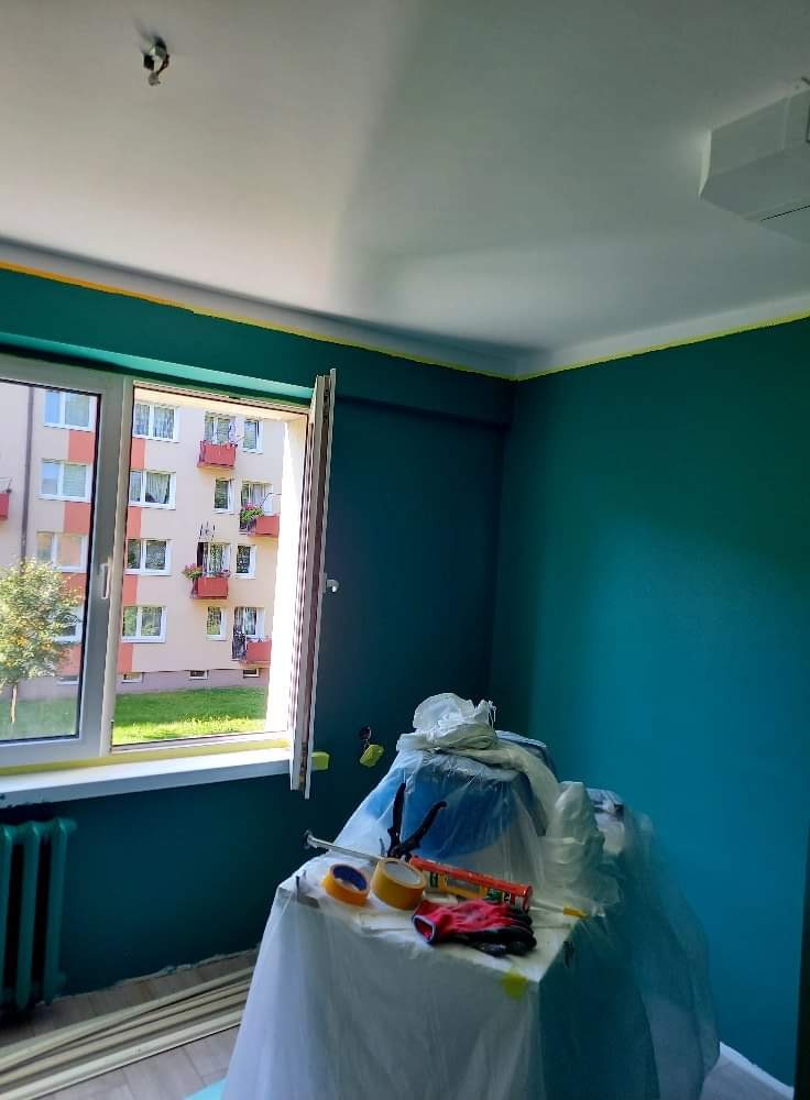 Pokój w trakcie malowania na ciemnozielono, z widocznymi narzędziami malarskimi na stole przykrytym folią, taśma malarska przy suficie i oknie, oraz widok na blok z balkonami za oknem.