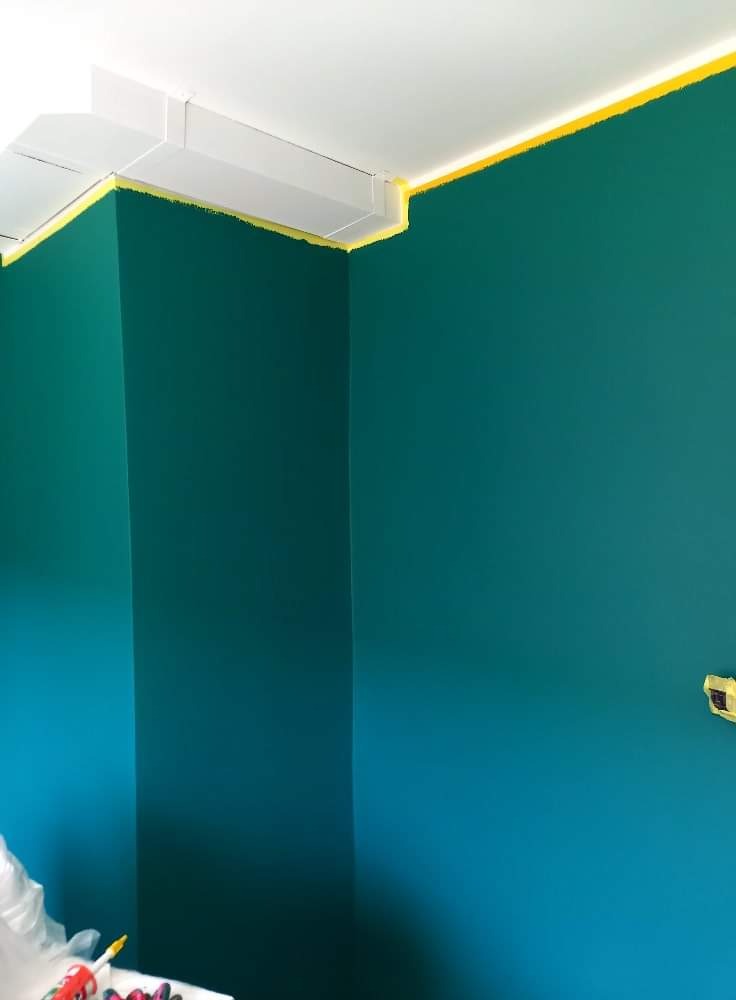 Ściana pomalowana na intensywny turkusowy kolor, zabezpieczona żółtą taśmą malarską przy białym suficie i narożniku, widoczne ślady farby na taśmie.