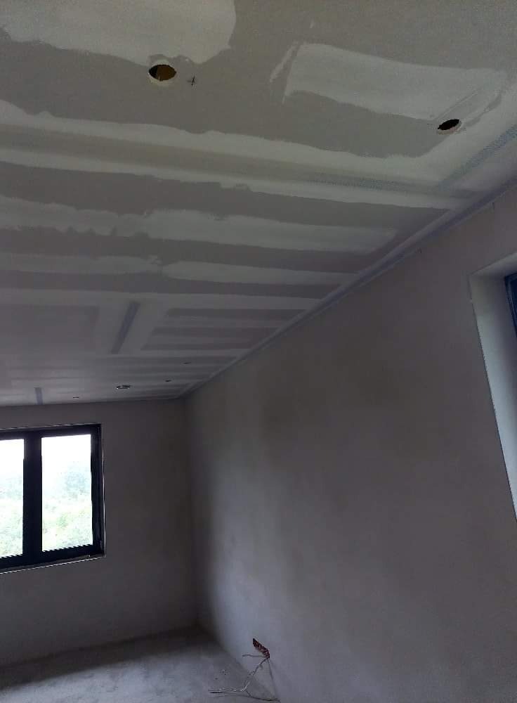 Surowy sufit z płyt kartonowo-gipsowych z widocznymi łączeniami i otworami na oświetlenie, ściany w stanie deweloperskim, okno z widokiem na zieleń.