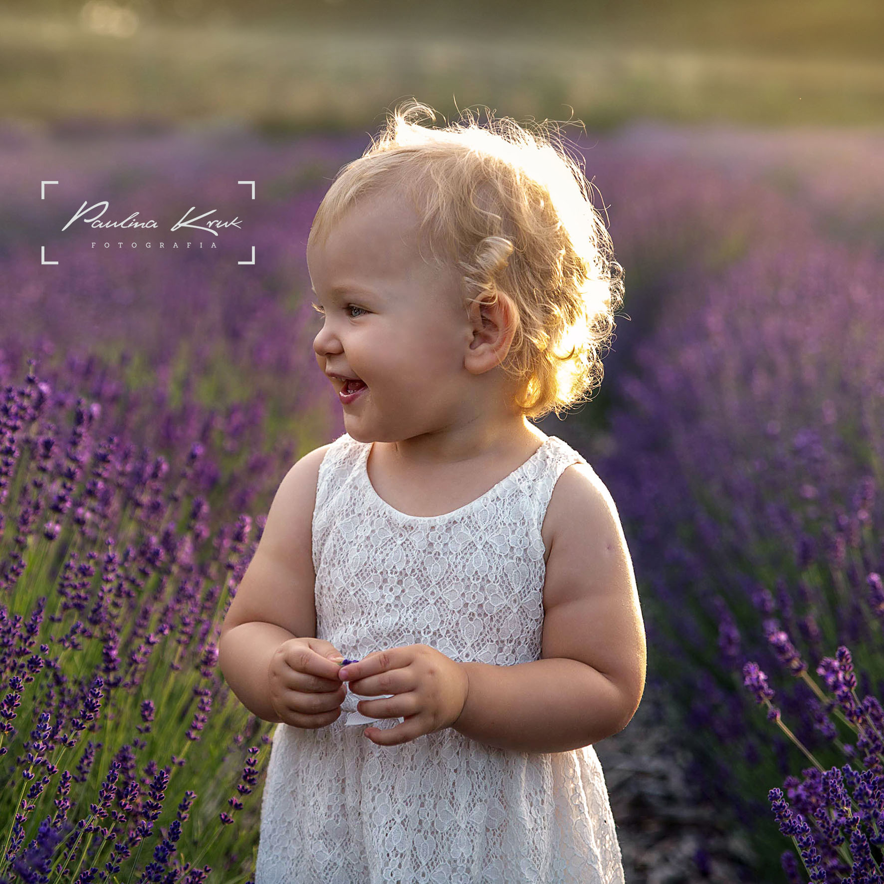 Uśmiechnięta dziewczynka w białej koronkowej sukience stoi na tle kwitnącej lawendy, oświetlona ciepłym, zachodzącym słońcem, z widocznym logo fotografa w lewym górnym rogu.