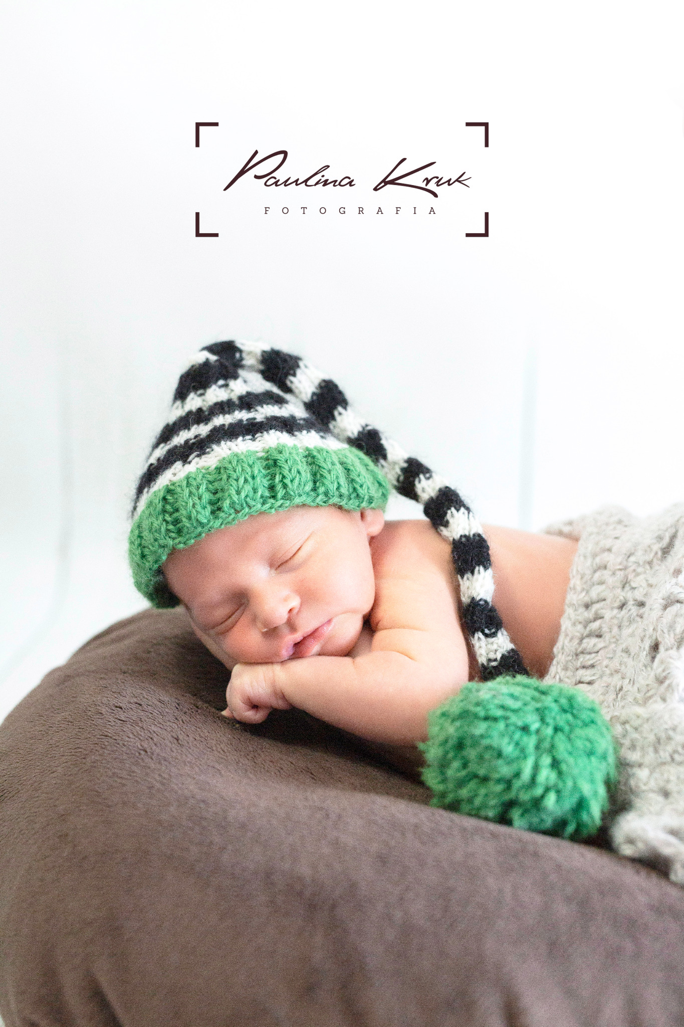 Śpiący noworodek w czapce w paski z zielonym pomponem, leżący na brązowej poduszce i przykryty szarym kocykiem, z logo fotografa w górnej części zdjęcia.