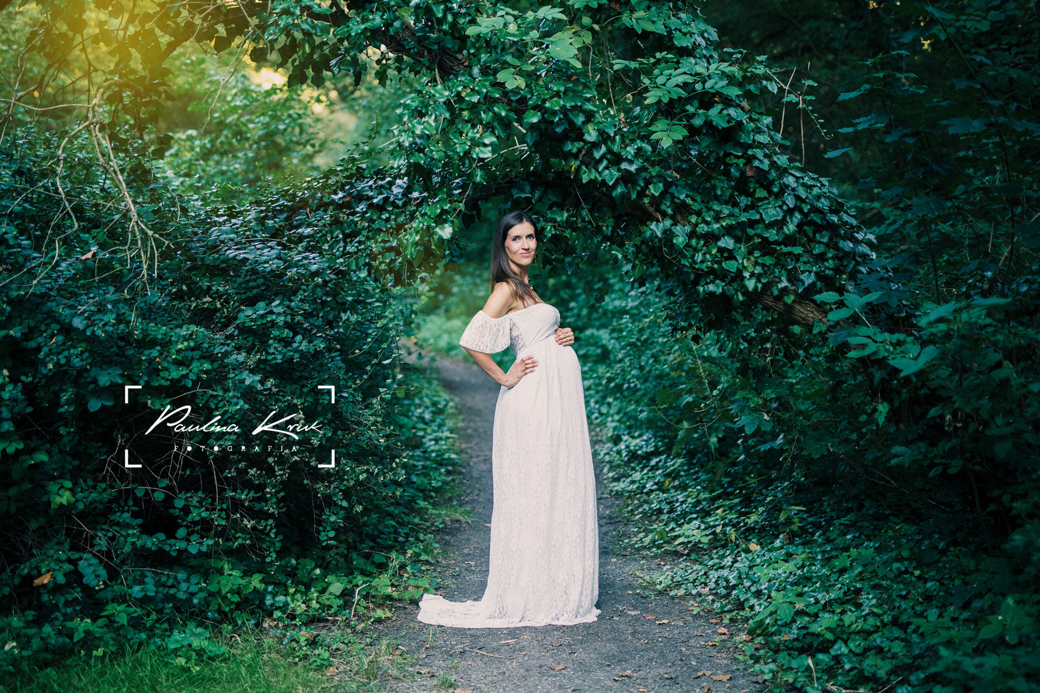 Kobieta w białej koronkowej sukni ciążowej pozuje na leśnej ścieżce, otoczona zielenią bluszczu i drzew, z delikatnym światłem słonecznym w tle.