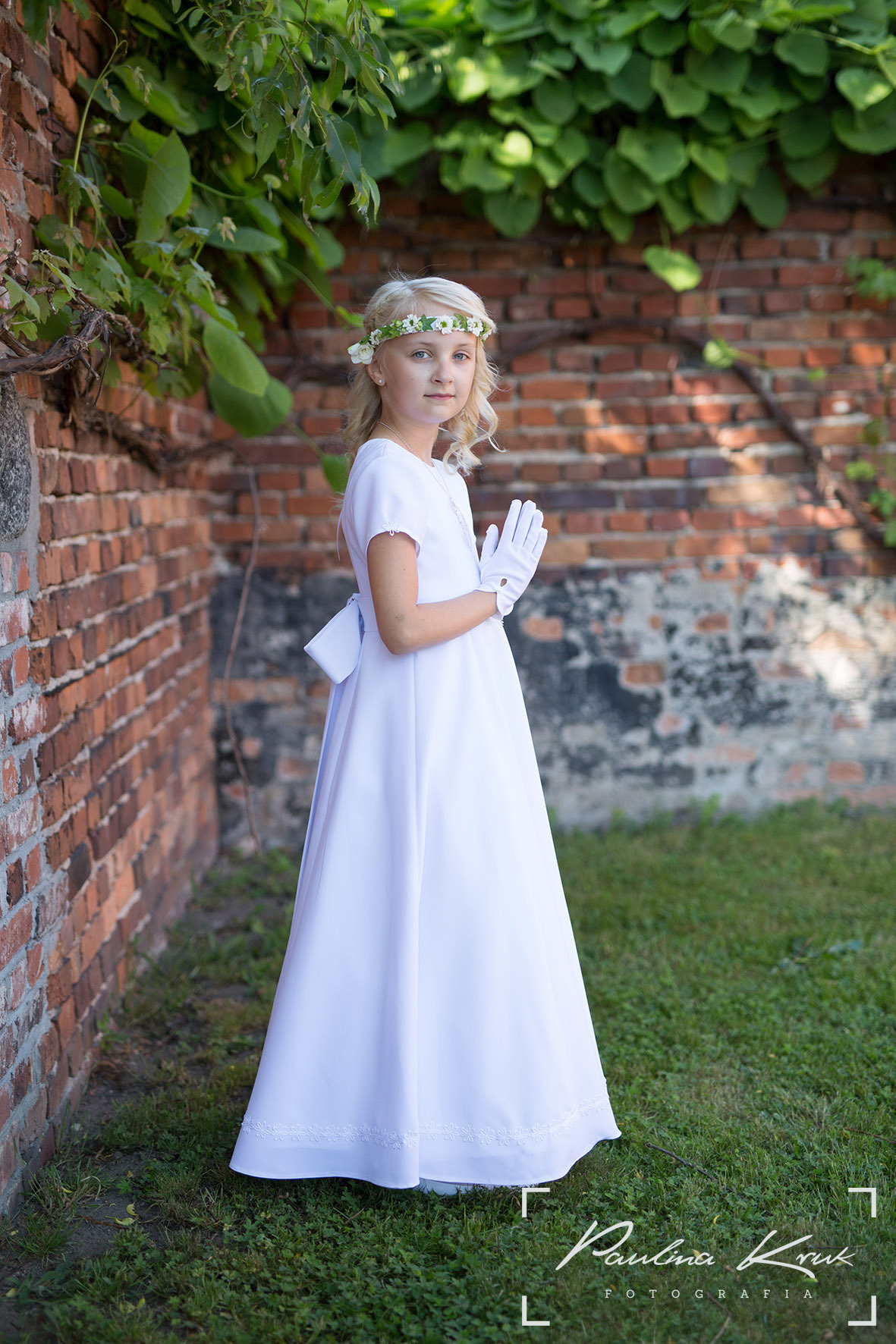 Dziewczynka w białej sukience komunijnej, z wiankiem na głowie i rękawiczkami, pozuje na tle ceglanej ściany porośniętej zielenią.