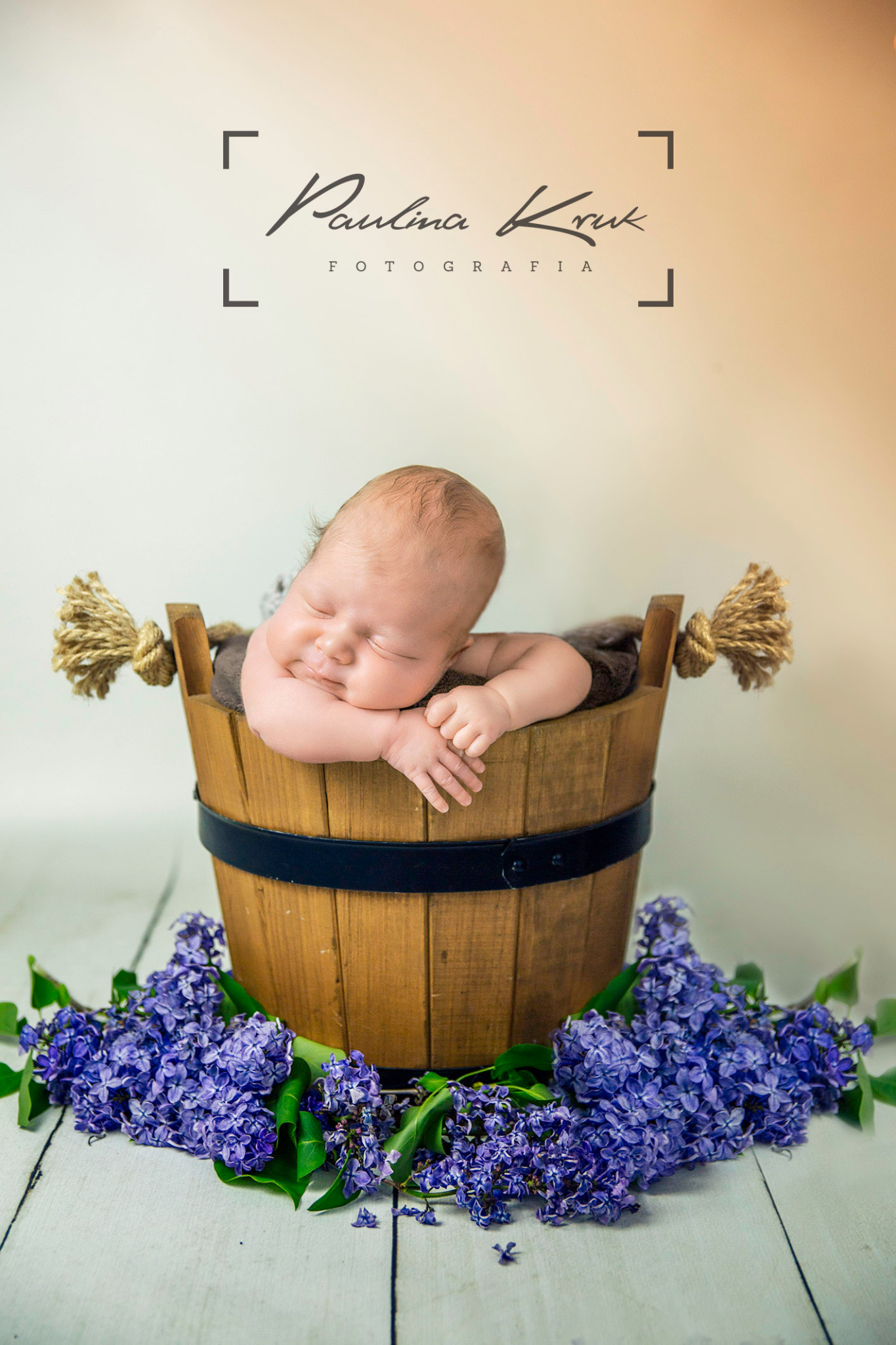 Śpiący noworodek ułożony w drewnianym wiaderku ozdobionym fioletowym bzem na białym, drewnianym tle, z logo fotografa w górnej części kadru.