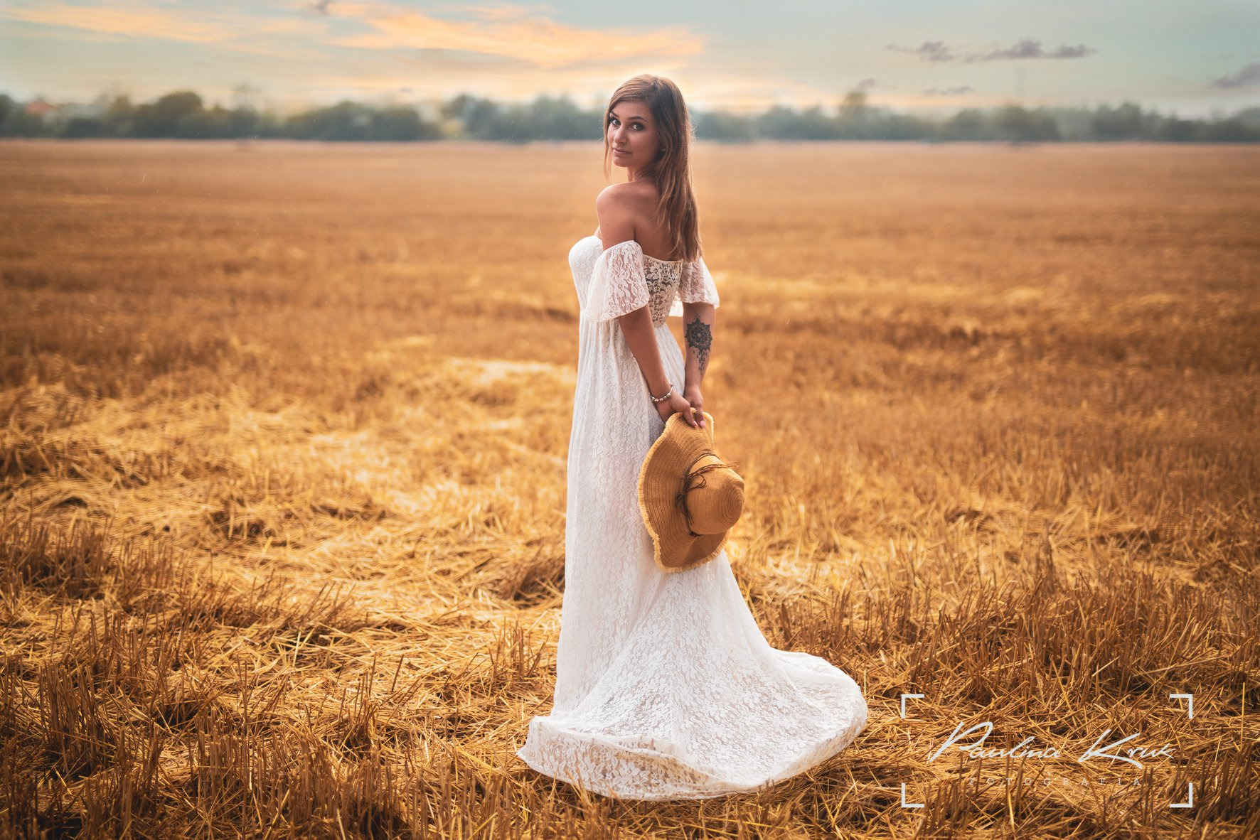 Kobieta w białej koronkowej sukni, z tatuażem na ramieniu, trzyma słomkowy kapelusz na tle pola po żniwach, spogląda przez ramię.