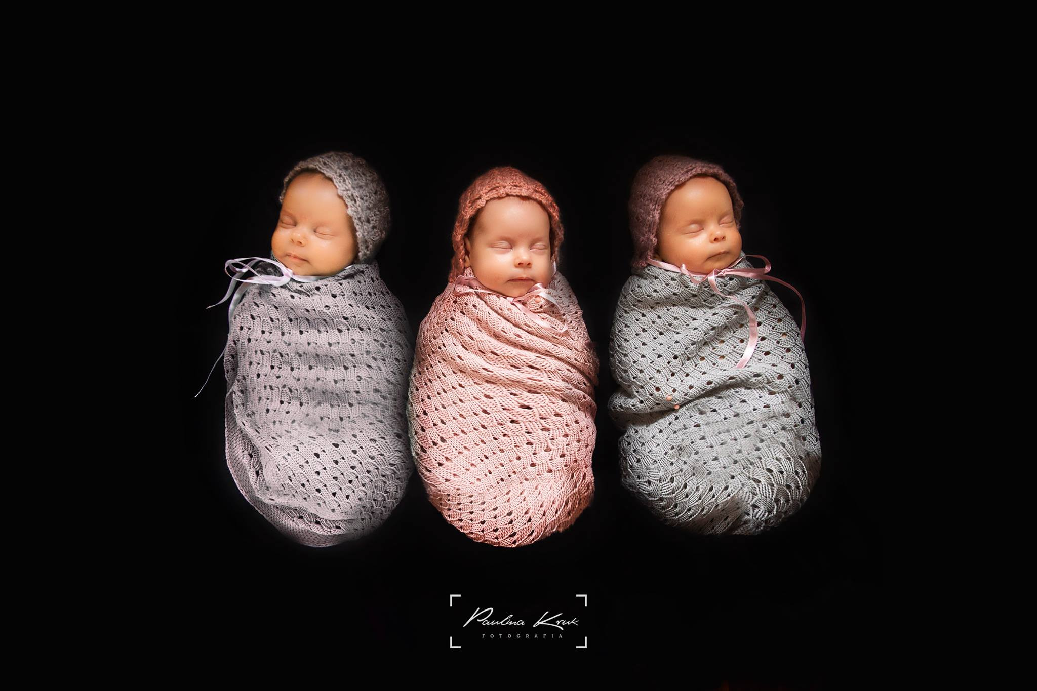 Trzy noworodki zawinięte w dziergane kocyki z kapturkami, śpiące obok siebie na czarnym tle, środkowy kocyk w odcieniu różu, dwa boczne w odcieniach szarości.