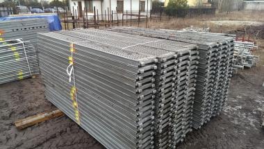 Sztaple metalowych podestów rusztowaniowych, zabezpieczone żółtą taśmą, składowane na placu budowy.
