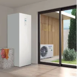Biała pompa ciepła Panasonic Aquarea w jasnym pomieszczeniu z drewnianą podłogą, widok na zewnętrzny moduł przez przesuwne drzwi tarasowe.