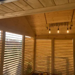 Drewniana konstrukcja z żaluzjowymi ścianami, oświetlona trzema żarówkami Edisona zawieszonymi na haku z grubym sznurem, widok na zewnątrz przez szpary w żaluzjach.