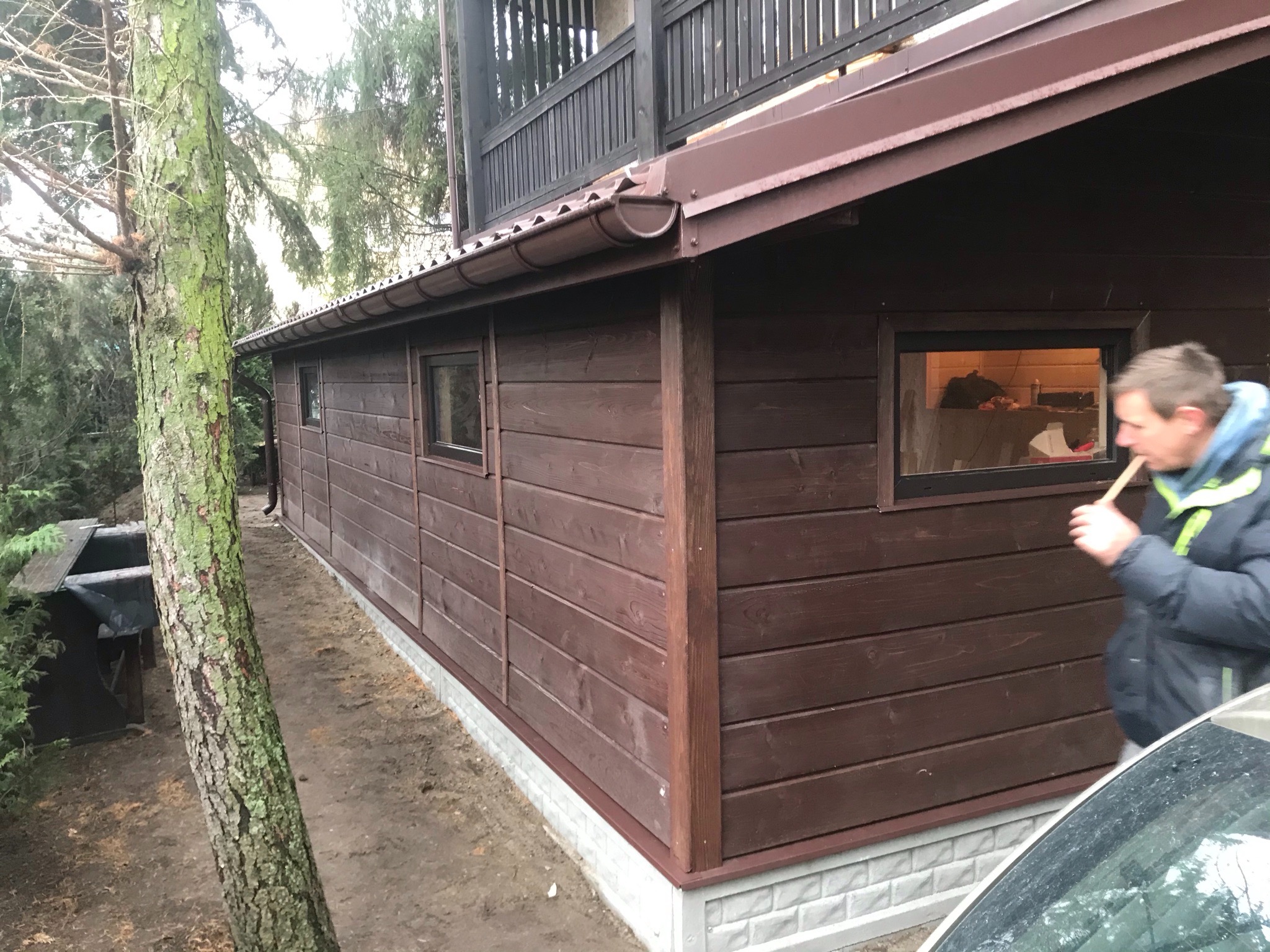 Brązowa drewniana konstrukcja z małymi oknami, widoczny fragment dachu i rynny, obok drzewo, w tle zieleń, w oknie widoczne przedmioty, obok konstrukcji stoi mężczyzna.