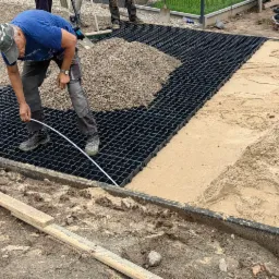 Pracownik układa czarną, plastikową kratkę drenażową na podsypce z piasku i żwiru, przygotowując podłoże pod nawierzchnię. Widoczne narzędzia i materiały budowlane.