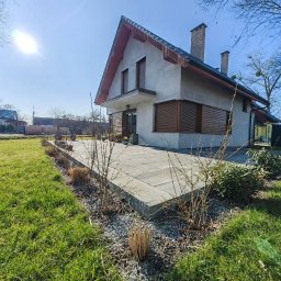 Metrikum Aleksander Atraszkiewicz - Nowoczesny taras z szarej kostki betonowej przy domu jednorodzinnym, z widocznym trawnikiem i elementami zieleni. Słoneczny dzień.